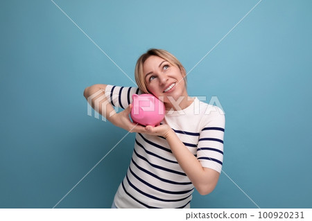 blond cheerful girl in casual attire carefully holds a piggy bank with savings on a blue background blond cheerful girl in casual attire carefully holds a piggy bank with savings on a blue background 100920231