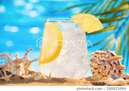 Refreshing crystal water with ice and lemon on a sea sandy beach with shells and palm leaves. Sunny day on vacation. 100921064