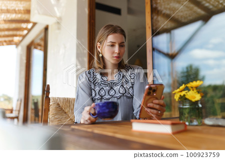 Young woman in ethnic outfit sitting at table with phone, working, having mug of morning coffee at home 100923759