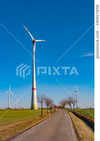 View over beautiful farm landscape with early spring agriculture field, wind turbines to produce green energy and a lonely road near Mittweida, Germany, at blue sunny sky. 100925698