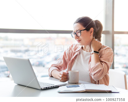 Woman winces from pain in her neck. Female office employee tries to stretch stiff neck muscles while working with laptop. Modern office at co-working center. Workplace for freelancers or students. Woman winces from pain in her neck. Female office employee tries to stretch stiff neck muscles while working with laptop. Modern office at co-working center. Workplace for freelancers or students. 100927358
