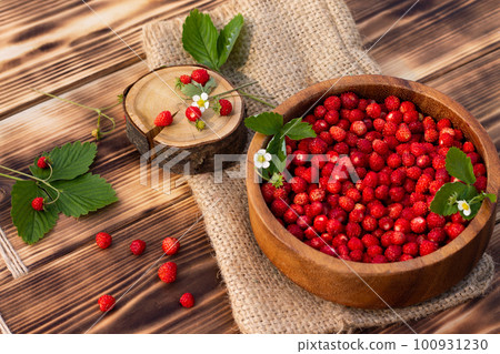 A wooden bowl of red ripe wild strawberries and flowers on old wooden surface. 100931230