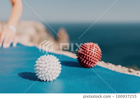 Woman sea pilates. Red and white massage balls are placed on a yoga mat, with the sea in the background. Healthy lifestyle, harmony and meditation. 100937255