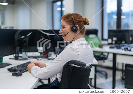 Red-haired caucasian woman in a wheelchair talking on a headset. Female call center worker smiling. 100942607