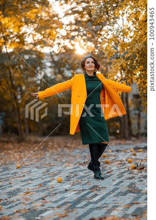 Beautiful woman walks outdoors in autumn. She is wearing a yellow coat and a green dress. Young woman enjoying the autumn weather. Autumn content. Beautiful woman walks outdoors in autumn. She is wearing a yellow coat and a green dress. Young woman enjoying the autumn weather. Autumn content. 100943465