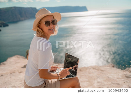 Freelance women sea working on the computer. Good looking middle aged woman typing on a laptop keyboard outdoors with a beautiful sea view. The concept of remote work. 100943490