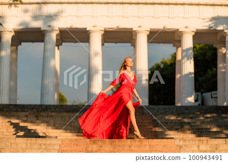 A woman in a long red dress against the backdrop of sunrise, bright golden light of the sun's rays. The concept of femininity, harmony. 100943491