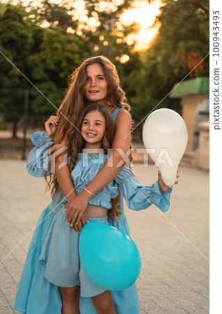 Mother daughter sunset. in blue dresses with flowing long hair against the backdrop of sunset. The woman hugs and presses the girl to her. They are looking at the camera. 100943493