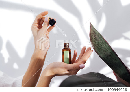 Woman in a white bathrobe takes care of her skin and moisturizes it her face with a serum 100944628