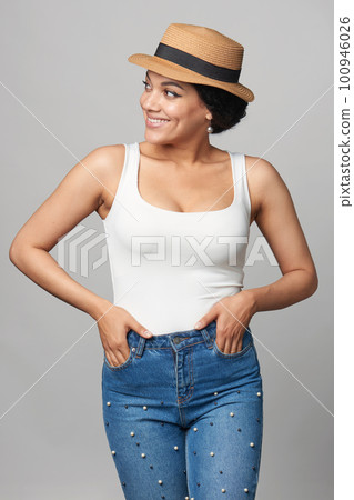 Happy mixed race woman in jeans and white tank top wearing canotier straw hat with hands in pockets 100946026