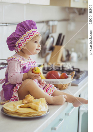 Adorable baby in apron and chef's cap making pancakes in the kitchen Adorable baby in apron and chef's cap making pancakes in the kitchen 100946803