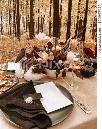 Table serving outdoors in the autumn forest. Black plate and napkin, card, fork, glass of champagne. Nuts and cheese on a plate for snacks. 100947015