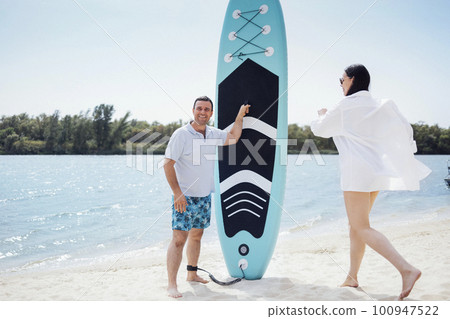 Surfers couple on the beach. Man stand with paddleboard and smiling. He waiting his partner. Young beautiful woman in sunglasses and white srirt running towards her boyfriend. 100947522