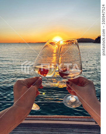 Female hands holding glasses of champagne on board the boat. Sunset sky and sea on the background. Making a celebratory toast with sparkling wine. Female hands holding glasses of champagne on board the boat. Sunset sky and sea on the background. Making a celebratory toast with sparkling wine. 100947524