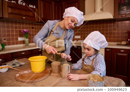 Authentic smiling happy woman, mother, confectioner removes raw dough from bowl and puts it into a baking dish, standing by kitchen table with her daughter, enjoying cooking Easter or Christmas cakes Authentic smiling happy woman, mother, confectioner removes raw dough from bowl and puts it into a baking dish, standing by kitchen table with her daughter, enjoying cooking Easter or Christmas cakes 100949623