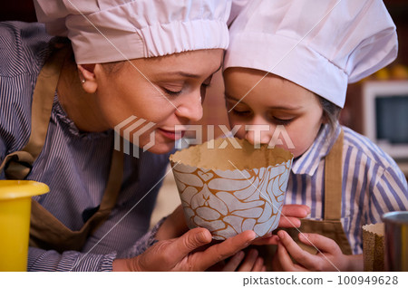 Close-up of a young multi-ethnic woman, a loving mother and her lovely little daughter, both of them wearing white chef's hat, sniffing dough while preparing Easter cake together in the home kitchen 100949628
