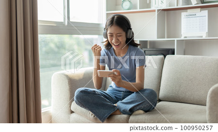 Asian woman playing games on mobile phone and wearing headphones having fun, Play games on the sofa in the living room on weekends, Resting at home, Comfort zone, Touch screen mobile phone. 100950257