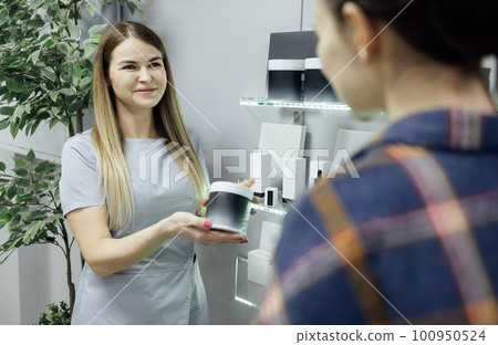 Smiling seller offers to choose the right cosmetics for female client. Plastic jar mockup in the hands of a seller woman. 100950524