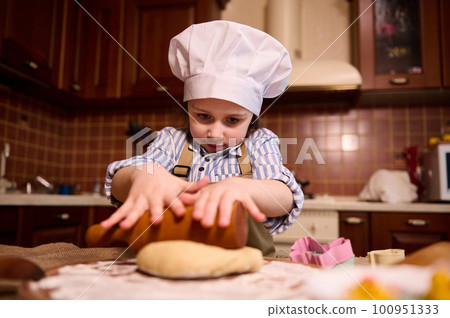 Caucasian lovely little toddler girl in white chef's hat, rolls out dough on floured wooden board, using rolling pin, preparing delicious gingerbread cookies for Easter holidays in the home kitchen 100951333