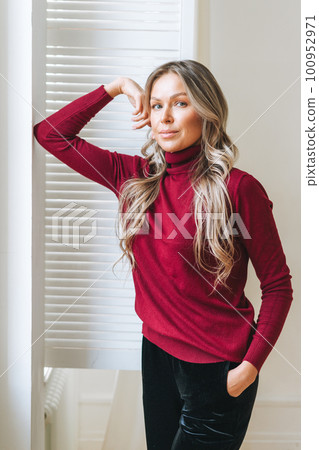 young woman with blonde long curly hair in bordo sweater in full length in bright interior young woman with blonde long curly hair in bordo sweater in full length in bright interior 100952971