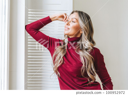 young woman in bordo sweater near window in bright interior, winter warm clothes 100952972