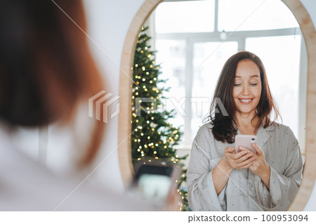 Young woman with dark hair looking at mirror and taking selfie on the Christmas morning in room  100953094