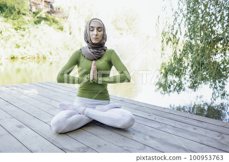 Muslim young girl in a hijab is doing yoga asana in the park. Teenage girl sits in a lotus position on pier by the lake or river. 100953763