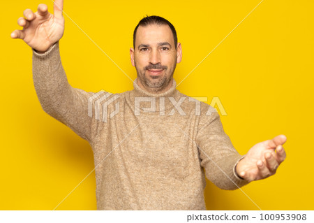 Bearded Hispanic man wearing a beige turtleneck in a friendly attitude looking at the camera smiling with open arms for a hug. Cheerful expression embracing happiness. Isolated on yellow background. Bearded Hispanic man wearing a beige turtleneck in a friendly attitude looking at the camera smiling with open arms for a hug. Cheerful expression embracing happiness. Isolated on yellow background. 100953908