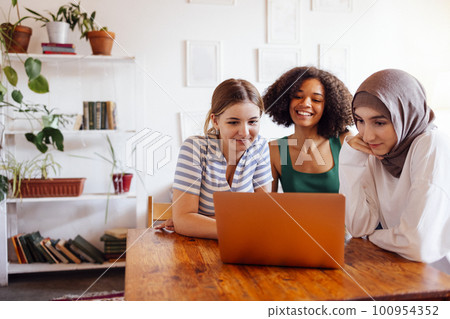 Three pretty female teenagers of different nationalities are looking at the laptop screen and laughing 100954352