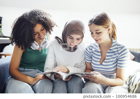 Three pretty female teenagers of different nationalities are reading a magazine and smiling 100954686