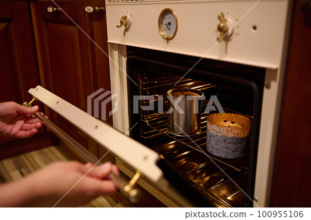 Close-up hands of a woman housewife, chef confectioner putting baking dishes with dough in the oven, cooking homemade delicious Easter cakes panettone in the home kitchen. Festive bakery and culinary Close-up hands of a woman housewife, chef confectioner putting baking dishes with dough in the oven, cooking homemade delicious Easter cakes panettone in the home kitchen. Festive bakery and culinary 100955106
