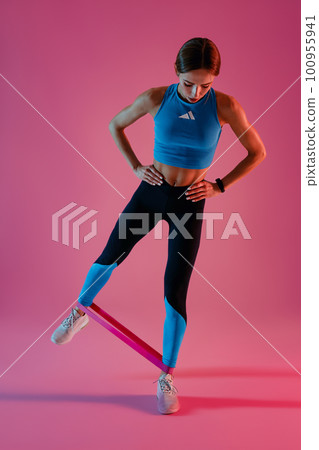 Athletic active woman standing and stretching elastic band with legs on studio background Athletic active woman standing and stretching elastic band with legs on studio background 100955941
