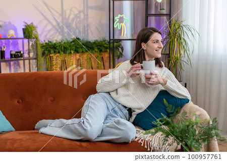 Attractive smiling young woman girl drinking a cup of coffee or herbal tea at home sofa in morning 100957761
