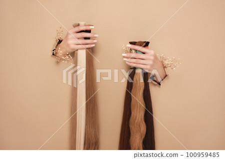 Hands with flowers hold strands of hair for extensions on a beige background. 100959485