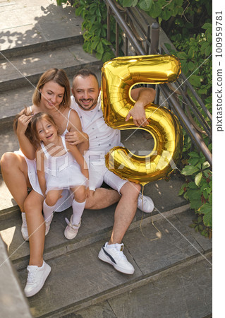 A cheerful, beautiful young family with a little daughter are sitting on the steps of the stairs in the courtyard with a large balloon in the shape of the number five. Child's birthday 100959781