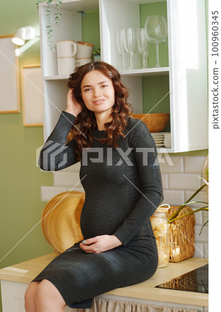 a slender pregnant woman in a black dress sits on a table in a home interior. a slender pregnant woman in a black dress sits on a table in a home interior. 100960345