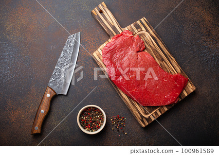 Raw uncooked top round beef steak on wooden cutting board with big kitchen knife and pepper on dark brown rustic stone background top view, cooking meat steak Raw uncooked top round beef steak on wooden cutting board with big kitchen knife and pepper on dark brown rustic stone background top view, cooking meat steak 100961589