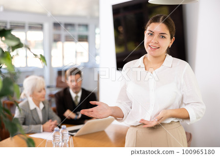 positive female colleague standing in meeting room and employees are talking behind her at table 100965180