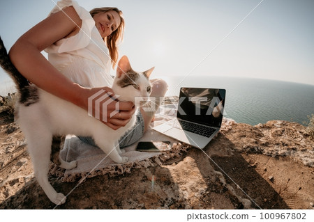 Woman sea laptop. Business woman petting cat and working on laptop by the sea. Close up on hands of pretty lady typing on computer outdoors summer day. Freelance, digital nomad and holidays concept. Woman sea laptop. Business woman petting cat and working on laptop by the sea. Close up on hands of pretty lady typing on computer outdoors summer day. Freelance, digital nomad and holidays concept. 100967802