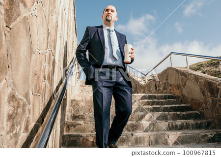 Confident middle age businessman with briefcase walking upstairs. Close-up of businessman wearing blue suit holding bag and coffee cup walking down the stairs Confident middle age businessman with briefcase walking upstairs. Close-up of businessman wearing blue suit holding bag and coffee cup walking down the stairs 100967815