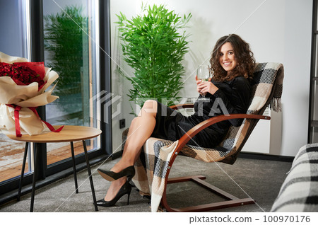 Curly young woman enjoying weekends inside contemporary barn house. Happy female tourist sitting on chair with wine and bouquet of roses in new cottage. Curly young woman enjoying weekends inside contemporary barn house. Happy female tourist sitting on chair with wine and bouquet of roses in new cottage. 100970176