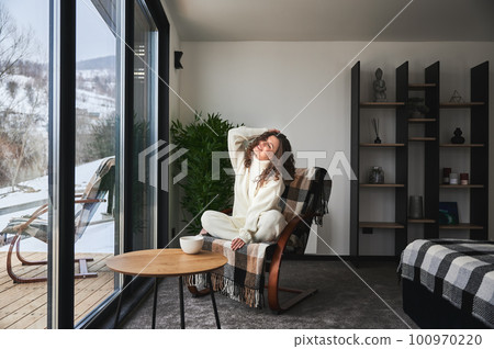 Curly young woman enjoying weekends inside contemporary barn house in the mountains. Happy female tourist sitting on chair, holding cup tea in new cottage with panoramic windows. Curly young woman enjoying weekends inside contemporary barn house in the mountains. Happy female tourist sitting on chair, holding cup tea in new cottage with panoramic windows. 100970220