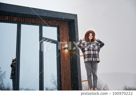 Curly young woman resting on terrace of modern barn house in the mountains. Happy female tourist enjoying winter holidays in new cottage. 100970222