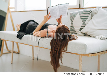 Beautiful young woman with dark long hair in black dress reading book in white interior Beautiful young woman with dark long hair in black dress reading book in white interior 100972094