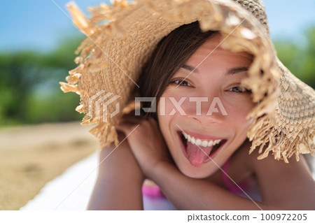 Happy funny face Asian young woman doing goofy facial expression sticking tongue out on summer vacation relax holiday sun tanning on beach having fun. 100972205