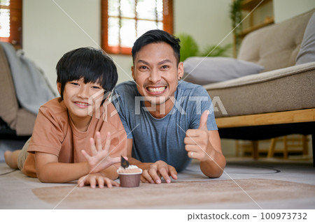 Happy Asian dad showing thumb up while laying on the living room floor with his son 100973732