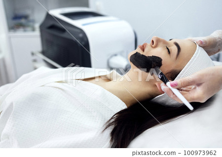 Female cosmetologist applying black mask on the face of a beautiful woman 100973962