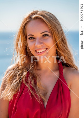 Smiling young woman in a red dress looks at the camera. A beautiful tanned girl enjoys her summer holidays at the sea. Portrait of a stylish carefree woman laughing at the ocean. Smiling young woman in a red dress looks at the camera. A beautiful tanned girl enjoys her summer holidays at the sea. Portrait of a stylish carefree woman laughing at the ocean. 100974193