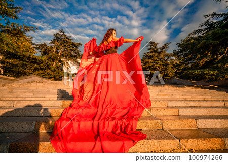 Sunrise red dress. A woman in a long red dress against the backdrop of sunrise, bright golden light of the sun's rays. The concept of femininity, harmony. 100974266