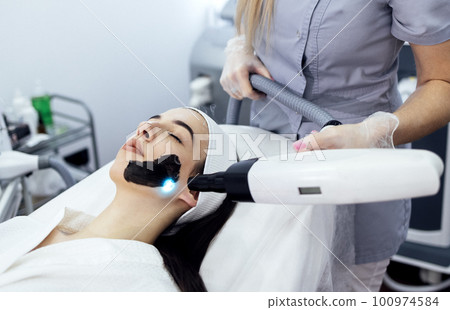 Beautician holding beauty laser near woman face covered with carbon mask Beautician holding beauty laser near woman face covered with carbon mask 100974584